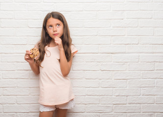 Young hispanic kid over white brick wall eating chocolate chips cooky serious face thinking about question, very confused idea