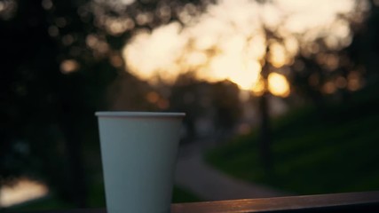 A white paper cup with coffee or tea stands on the railing against the backdrop of the city park at sunset. A hot drink raises steam. - Powered by Adobe