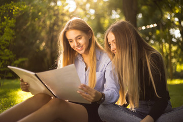 Young beautiful ladies students sitting in the park outdoors on grass holding copybook doing homework.