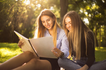 Young beautiful ladies students sitting in the park outdoors on grass holding copybook doing homework.