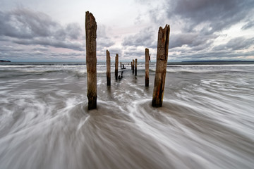 Wooden poles on Baltic Sea beach in high contrast evening light with clouds in windy weather, water movement in long exposure - Location: Baltic Sea, Rügen Island