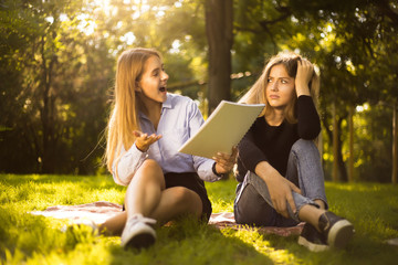 Excited girl try to force her displeased confused friend sister to education students sitting in the park outdoors on grass holding copybook doing homework.