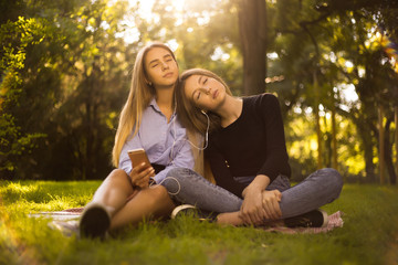 Obraz premium Sisters students sitting in the park outdoors on grass sleeping while have a rest meditate listening music with earphones.