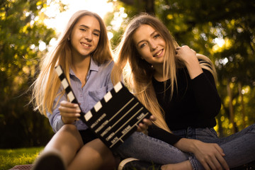 Happy girls sisters students sitting in the park outdoors on grass have a rest holding film making clapperboard.