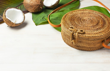 Fashionable handmade natural organic round rattan bag, fresh coconut and tropical leaves on light wooden background flat lay with copy space. Trendy bamboo bag Ecobags from Bali Summer fashion concept