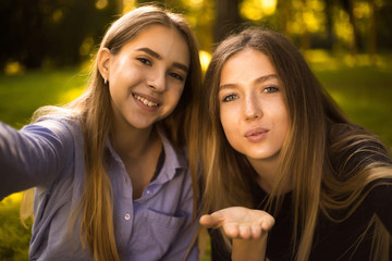 Obraz premium Happy laughing girls sisters students sitting in the park outdoors on grass have a rest take a selfie by camera blowing kisses.