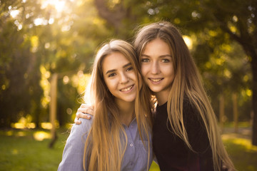 Fototapeta premium Happy laughing girls sisters students standing in the park outdoors on grass have a rest looking camera hugging.