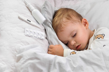 Sick little boy sleeping in white bed with a nasal spray, thermometer and antipyretic tablets on...