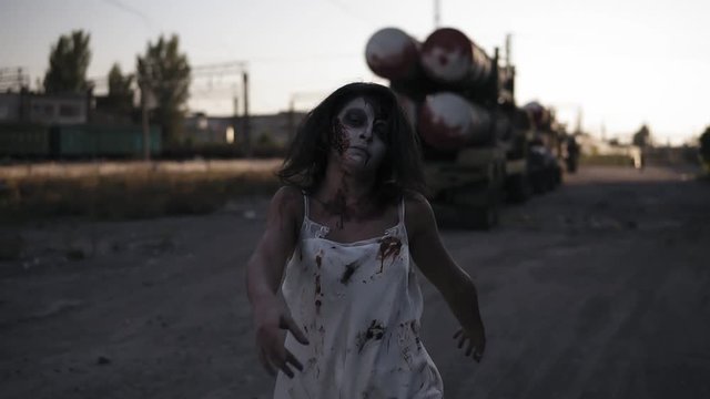 Creepy zombie woman with wounded face walking outdoors with an industrial abandoned place on the background. Halloween, filming, creepy concept