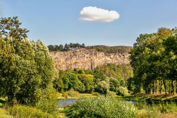 Fototapeta na wymiar View of valley with river and high brown pink steep limestone rock, trees lined on the top of the rock, white cloud in the middle of blue sky, bright fall sunny day, bushes in foreground