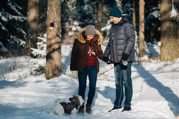 Fototapeta na wymiar Cute young couple having fun in winter forest with their pretty little white dog.