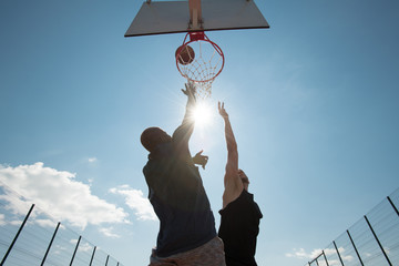 Low angle view of two young men playing basketball and jumping by hoop against blue sky, copy space - Powered by Adobe