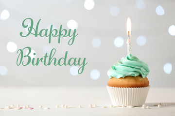 Delicious cupcake with burning candle and greeting HAPPY BIRTHDAY on blurred lights background