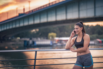 Fototapeta premium Happy young sporty fitness woman having a chat over her cell phone, sunset view in an urban outdoor area
