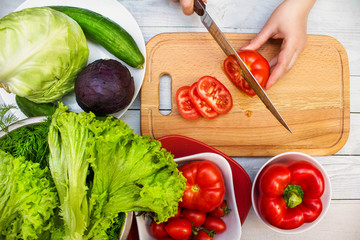 Hands of a cook cutting tomatoes for making a fresh vegetable salad. Set of delicious healthy food products: tomatoes, cucumbers, sweet pepper, cabbage and lettuce salad.