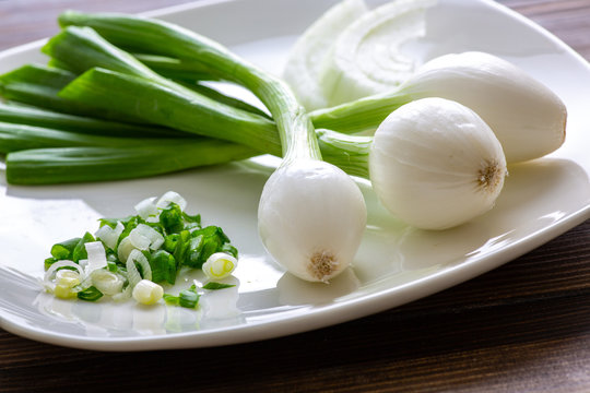 Three Large Mexican Onions Which Are Green Onions That Have Been Allowed To Grow Bigger On A White Plate With Some Smaller Green Onions Sliced Next To It On A Kitchen Table.