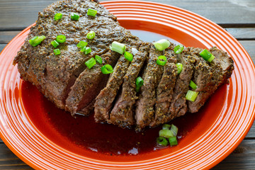 Large tri-tip church of beef just off the grill cooked medium well on a red plate with green onion garnish in its own juice on the kitchen table