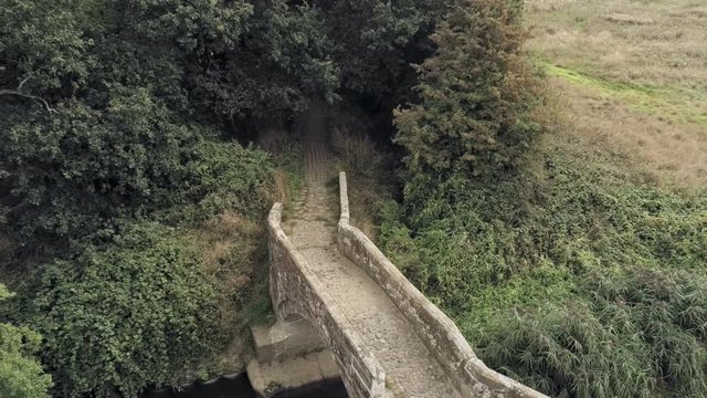 Aerial view, down move revealing old stone roman bridge and grass, overgrown water stream in english countryside.