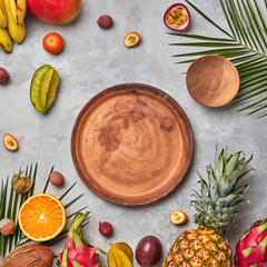 Various juicy exotic fruits, coconut, lychees, carom, pineapple, palm leaves and empty brown wooden plates on a gray concrete background with space for text. Flat lay