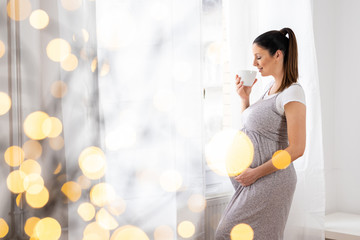 A young pregnant woman standing next to the window in an elegant grey dress and drinking a coffee from a mug with soft bokeh lights.