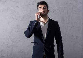 A serious handsome young businessman in a black suit talking on his phone in front of a grey wall in a studio.