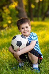 Portrait of a cute little boy with football sitting on grass in the park