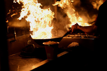 The cook in the kitchen holds a wok with an open flame in his hands - 224103716
