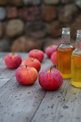 Apple cider or juice beverage in two glass bottles and red ripe fresh apples on grey natural wooden table background with rain drops and copy space for text.