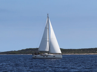 A modern cruise sailing yacht with a Bermuda sloop-type rig goes past the green coast of the Croatian Riviera on a sunny summer day. Adriatic Sea of the Mediterranean region. District of Dalmatia