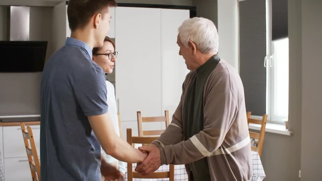 Medium shot of two volunteers hugging and shaking hands with elderly man and talking to him when visiting him at home