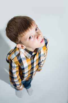 Little boy in checkered shirt and jeans on white background stands, looks up thoughtfully and slyly