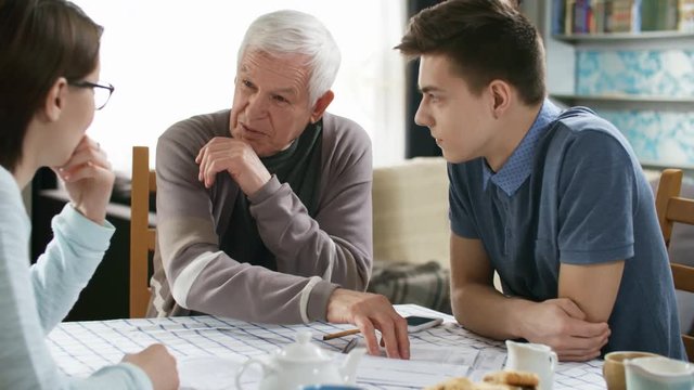 Medium shot of retired man sitting at table and telling something to two young caregivers or grandchildren