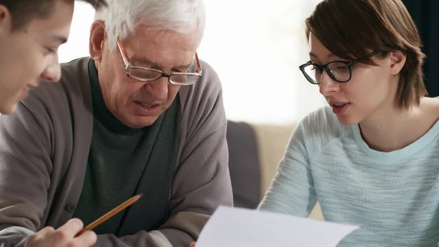 Medium shot of two caregivers sitting at kitchen table and helping retired man in glasses understand household bills