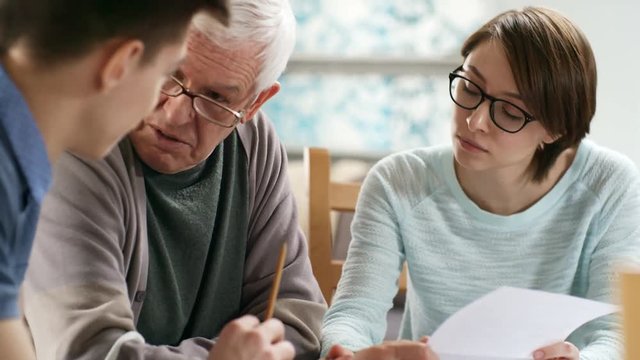 Medium shot of retired man in glasses trying to understand household bills and talking to two caregivers sitting next to him