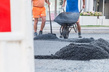 Straßenarbeiter verteilen den heißen Asphalt mit der Schaufel und Schubkarre auf der Straße,...