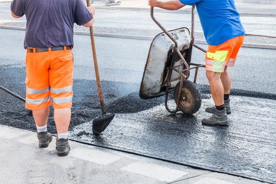 Stra&szlig;enarbeiter verteilen den hei&szlig;en Asphalt mit der Schaufel und Schubkarre auf der Stra&szlig;e, Deutschland