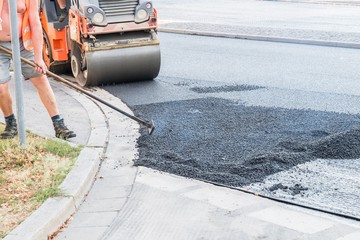 Straßenarbeiter verteilen den heißen Asphalt mit der Schaufel und walzen diesen auf der Straße,...