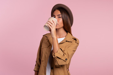 Young woman drinks coffee in the morning, wearing hat and autumn coat isolated over pink background