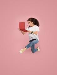 Gadget in modern life. Jump of young woman over pink studio background using laptop or tablet gadget while jumping. Runnin surprised girl in motion or movement. Human emotions and facial expressions