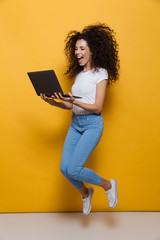 Full length photo of funny woman 20s wearing casual clothes smiling and jumping while holding black laptop, isolated over yellow background