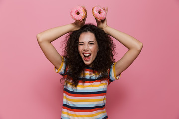 Photo of beautiful woman 20s with curly hair having fun and holding donuts, isolated over pink background