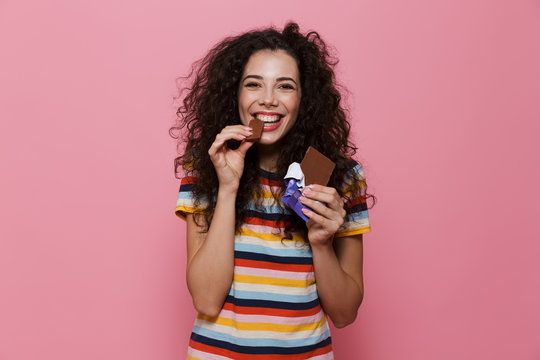 Photo of lovely woman 20s with curly hair eating chocolate bar, isolated over pink background