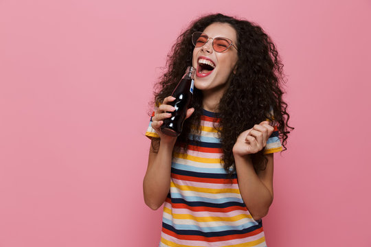 Photo of happy woman 20s with curly hair drinking soda from glass bottle, isolated over pink background