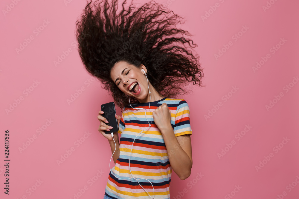 Poster Image of cute woman 20s with shaking hair singing while holding smartphone and listening to music via headphones, isolated over pink background