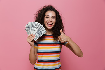 Image of young woman 20s with curly hair holding fan of dollar money, isolated over pink background