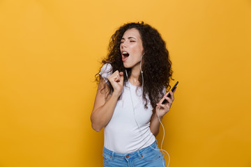 Obraz premium Image of happy woman 20s with curly hair singing while holding smartphone and listening to music via headphones, isolated over yellow background