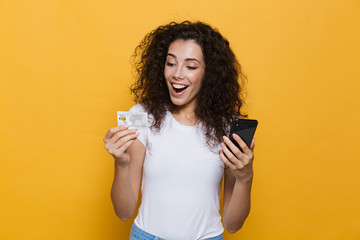 Photo of beautiful woman 20s wearing casual clothes holding mobile phone and credit card, isolated over yellow background