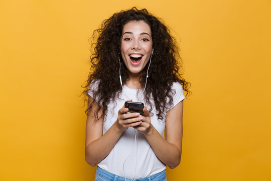 Image of excited woman 20s with curly hair holding smartphone and listening to music via headphones, isolated over yellow background