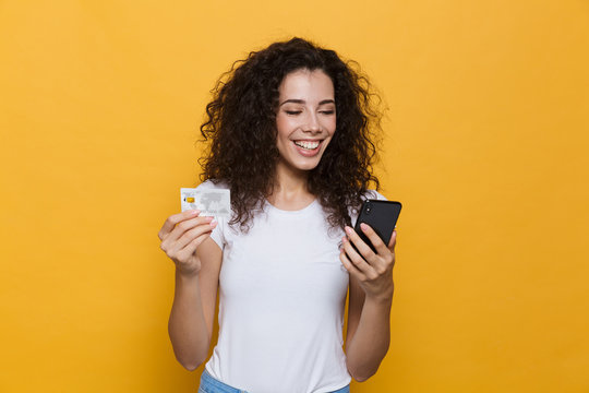 Photo of pretty woman 20s wearing casual clothes holding mobile phone and credit card, isolated over yellow background