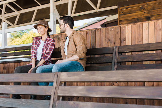 cheerful cowboy and cowgirl in casual clothes sitting on bench at ranch stadium